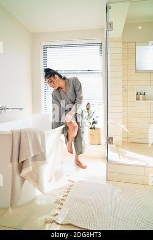 Woman in bathrobe preparing soaking tub for bath in sunny bathroom Stock Photo