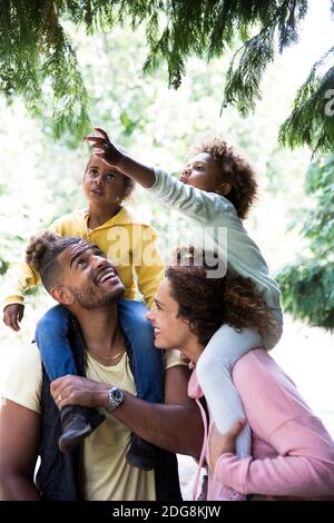 Happy parents carrying daughters on shoulders below trees in park Stock Photo