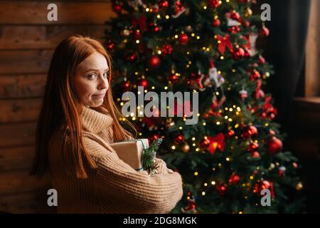 Side view of flirty redhead young woman holding Christmas gift box on background of xmas tree. Stock Photo