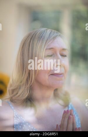 Serene senior woman praying with hands clasped at window Stock Photo