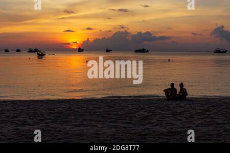 Koh Tao, Thailand: April 23 2019: island village boat pier, evening sunset time at beach, sitting couple on white sand coastline enjoy view Stock Photo