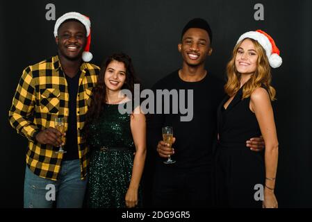 Waist up of four friends in Santa hats having fun and drinking champagne during New Year party, isolated on black background. Cheerful friends toastin Stock Photo