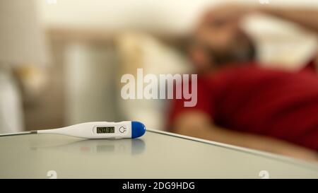 High fever temperature display on digital thermometer and infected ill man staying in bed at home. Pandemic stay safe virus protection equipment conce Stock Photo