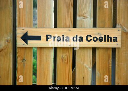 Wooden sign installed on a wooden fence showing direction towards Praia da Coelha or Rabbit Beach, a small beach surrounded by rock formations Stock Photo