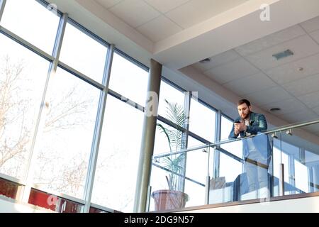 Smart beard Caucasian man is chatting texting on smartphone in urban modern coworking office on glass balcony near panoramic window. Businessman Stock Photo