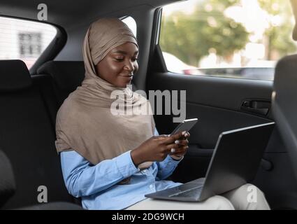 Black islamic businesswoman using smartphone and laptop, going to office by car Stock Photo