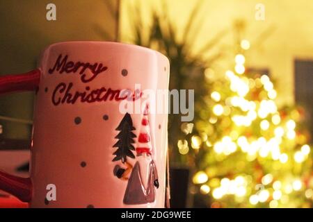 Close up hand painted red and white Christmas mug with blurred lit up Christmas tree in the background, out of focus lights, in Manchester, England