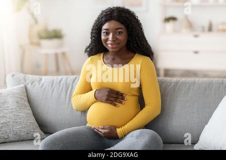 Beautiful black pregnant woman sitting on couch at home Stock Photo