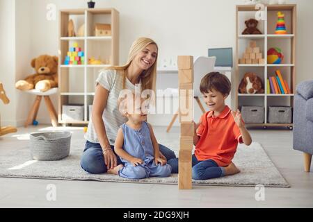 Mom with two small children have fun playing assembling a tower of wooden eco cubes at home. Stock Photo