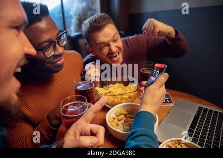 Happiness. Excited fans in bar with beer and mobile app for betting, score on their devices. Screen with match results, emotional friends cheering. Gambling, sport, finance, modern techn concept. Stock Photo