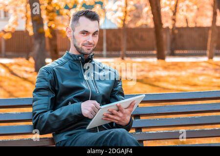 Handsome Young Man Sitting on the Bench and Using Big White Tablet PC at the Beautiful Autumn Park Stock Photo