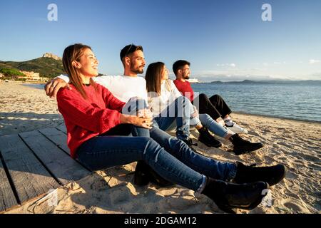 Four millennial friends sitting on a boardwalk of a beach in a sea resort looking at the sun goes down - Two young heterosexual couple in winter vacat Stock Photo