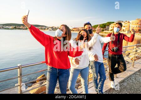 Group of friends wearing Coronavirus face protection mask making a selfie in row showing thumbs up in sign of victory on a sea winter vacation - Cauca Stock Photo