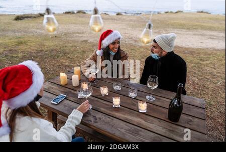 Three young beautiful people in Santa Claus hat sitting outdoor drinking champagne celebrating Christmas and New Year event in sea resort winter vacat Stock Photo