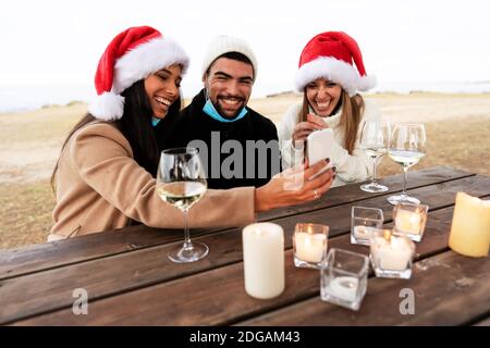Three happy young mixed-race friends sitting outdoor in Santa Claus hat smiling making selfie with lowered Coronavirus safe protection mask with to ce Stock Photo