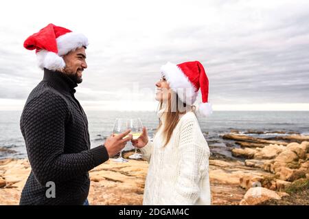Funny romance scene of a beautiful happy young couple celebrating Christmas standing outdoor face to face wearing Santa Claus hat toasting with champa Stock Photo
