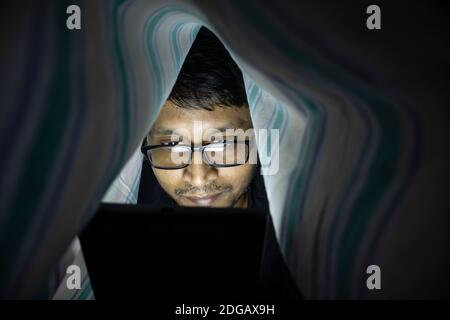 a middle aged Indian man looking at tablet or laptop screen at midnight secretly Stock Photo