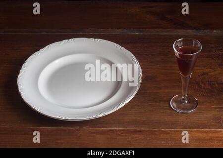 A shot glass of brown liquor next to an empty antique porcelain plate on a rustic chestnut wood tray. Stock Photo