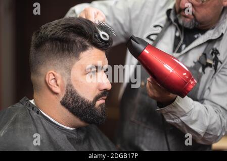 Close up view of master barber doing hairstyle with hairdryer at barber shop. Stock Photo