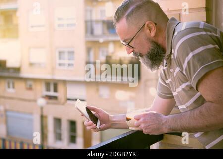 bearded man with glasses leaning on the railing of the balcony of the house at sunset having a coffee and using the smart-phone Stock Photo