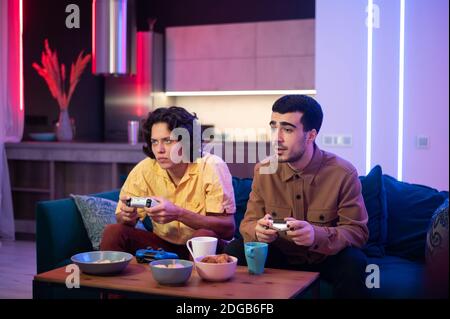 Smiling diverse male friends with gamepads playing video game at home. Portrait of latin and caucasian hipster guys playing game with console relaxing Stock Photo