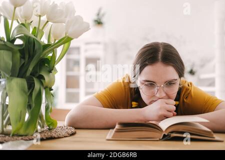Young beautiful woman student in orange T-shirt and glasses reads book sitting at the table. Preparing for exams, studying at home on self-isolation Stock Photo