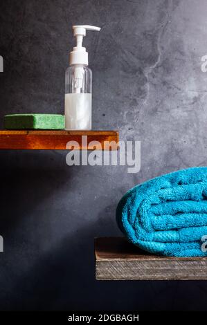 A turquoise towel, a soap dispenser, and a green bar of olive soap on wooden shelves against a gray background Stock Photo