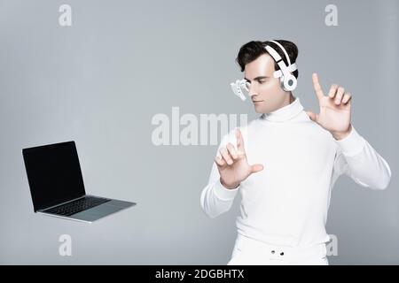 Cyborg in headphones touching something and looking at laptop with blank screen in air isolated on grey Stock Photo