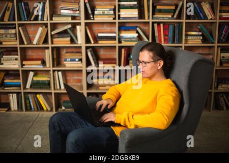 Smart adult male in casual clothes and glasses resting in comfortable armchair and browsing laptop against bookshelf in cozy library Stock Photo