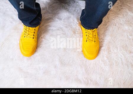 From above crop person holding legs in jeans and bright yellow sneakers on fluffy soft carpet at home Stock Photo