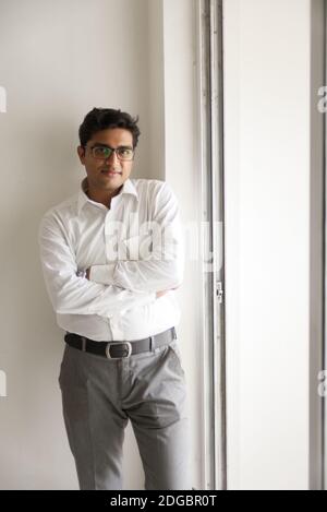 Portrait of an Indian Bengali tall, dark, handsome brunette young man in office wear is standing in front of a glass window in a corporate office/bpo Stock Photo