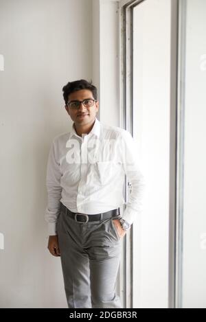 Portrait of an Indian Bengali tall, dark, handsome brunette young man in office wear is standing in front of a glass window in a corporate office/bpo Stock Photo