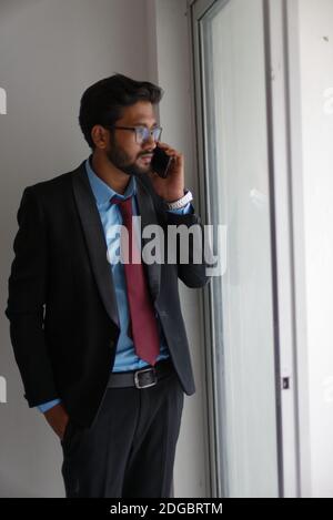 Portrait of an Indian Bengali tall, dark, handsome brunette young man in office wear is standing in front of a glass window in a corporate office/bpo Stock Photo