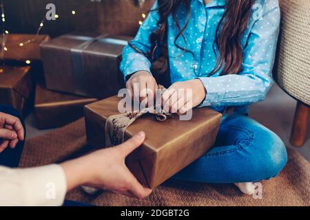 Merry Christmas and Happy New Year. Mom gives daughter present by tree at home. Girl unwraps gift box. Close up Stock Photo