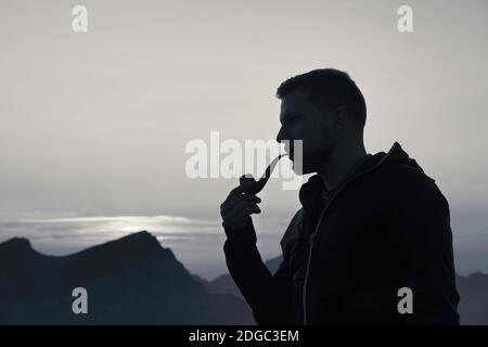 Young sporty attractive man smokes a pipe on mountain at sunset, enjoying the evening mood Stock Photo