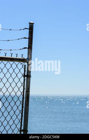 Edge of chain link fence and barbed wires against water and clear blue sky. Stock Photo