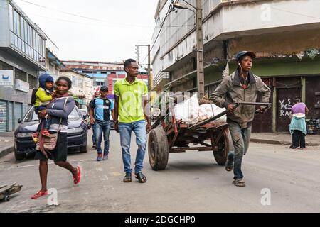 Antananarivo, Madagascar - April 24, 2019: Unknown Malagasy man pulling cart with white bags, woman with child on her hands walking near, more locals Stock Photo