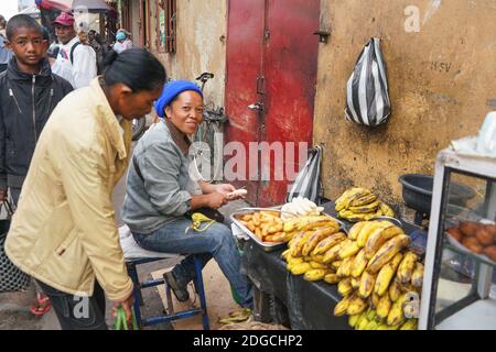 Antananarivo, Madagascar - April 24, 2019: Unknown Malagasy woman preparing food from bananas on street, more locals walking in background. Typical sc Stock Photo