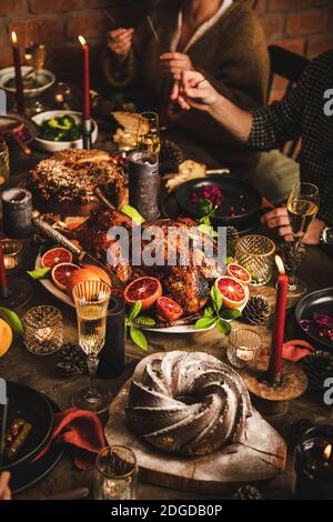 Family celebrating Christmas over festive table with roasted chicken in oranges, chocolate bundt cake, champagne and candles, red brick wall at background. Christmas, New Year holiday table setting Stock Photo