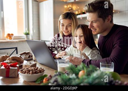 Happy parents with excited kid daughter using laptop sitting at Christmas table. Stock Photo