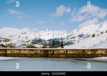 A group of hikers on snowshoes crossing a low dam by a frozen lake against snowcapped mountains, Panticosa, Aragon, Spain Stock Photo