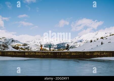 A group of hikers on snowshoes crossing a low dam by a frozen lake against snowcapped mountains, Panticosa, Aragon, Spain Stock Photo