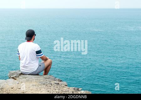 Young man with cap and sunglasses sitting on a cliff in front of the sea. Stock Photo