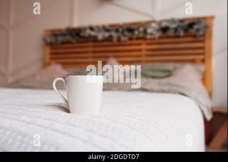 clear white ceramic mug on cozy wooden bed in christmas decor with bedspread of pastel colors, pillows of powdery shades and blanket of delicate color Stock Photo