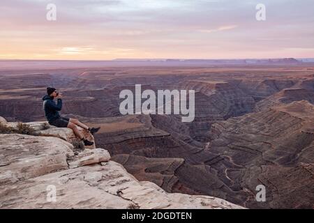 Man drinks coffee sitting on edge of cliff, Mexican Hat, Utah Stock Photo