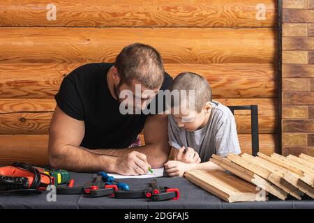 Dad and son are drawing on paper, planning how to build a bird house, tools and a beam on the table in the workshop. Carpentry training concept for ki Stock Photo
