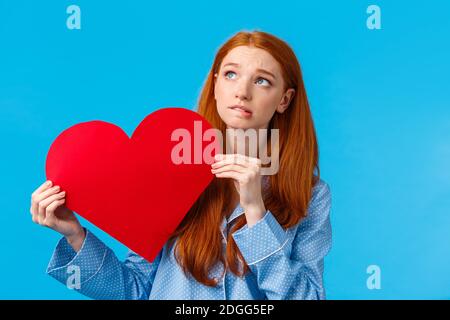 Shy and indecisive cute girl scared confess love, tell how she feels. Worried cute redhead teenager holding romantic heart card Stock Photo
