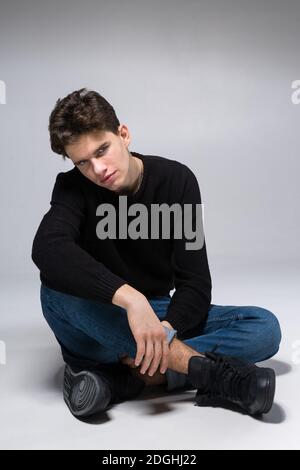Stylish handsome man posing sitting on floor. Handsome Guy. cool fashion male model sitting on grey background and looking at th Stock Photo