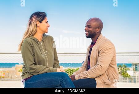 young interracial couple sitting at seaside on a bench looking at each other and interactig.happy people outside on autumn holidays.fiends on leasure Stock Photo
