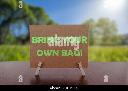 Bring your own bag text on card on the table with sunny green park background. Ecology concept, recycle, reuse, reduce waste. Stock Photo